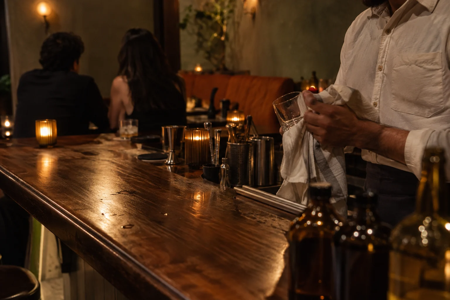 A bartender drying a glass as guests sit at the walnut counter